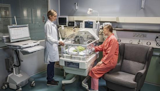 NICU patient in an incubator with a clinician and the mother's baby next to the patient